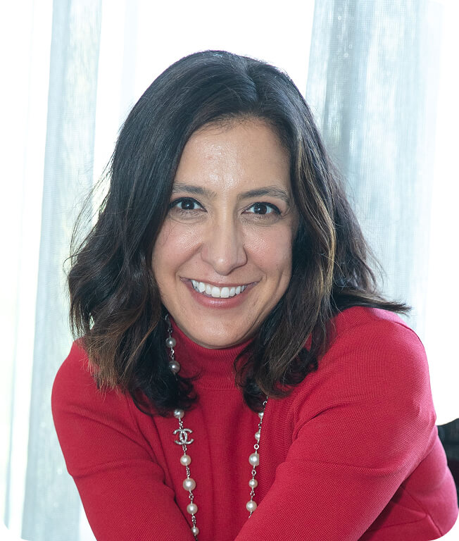 Professional portrait of founder Laura wearing a red sweater and pearl necklace, smiling against a bright, soft-focus background.